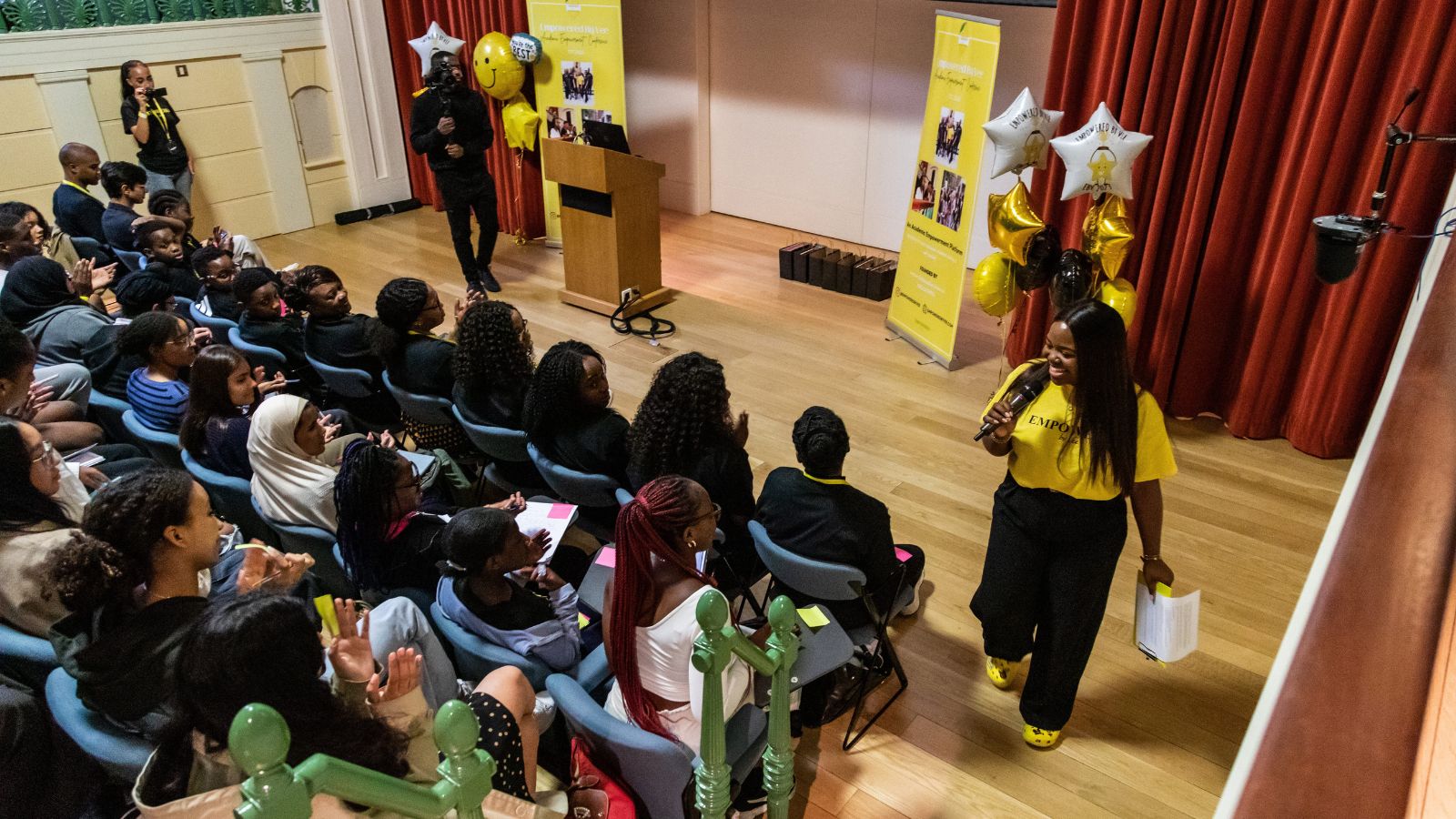 A group of young people in a lecture theatre, with Vee Kathivu wearing a yellow tshirt and holding a microphone