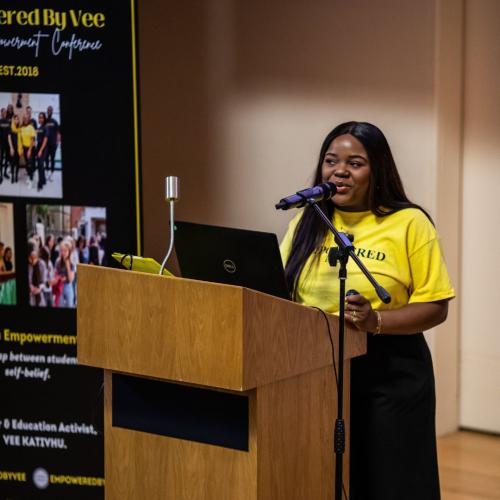 Vee Kathivu standing at a podium wearing a bright yellow t-shirt