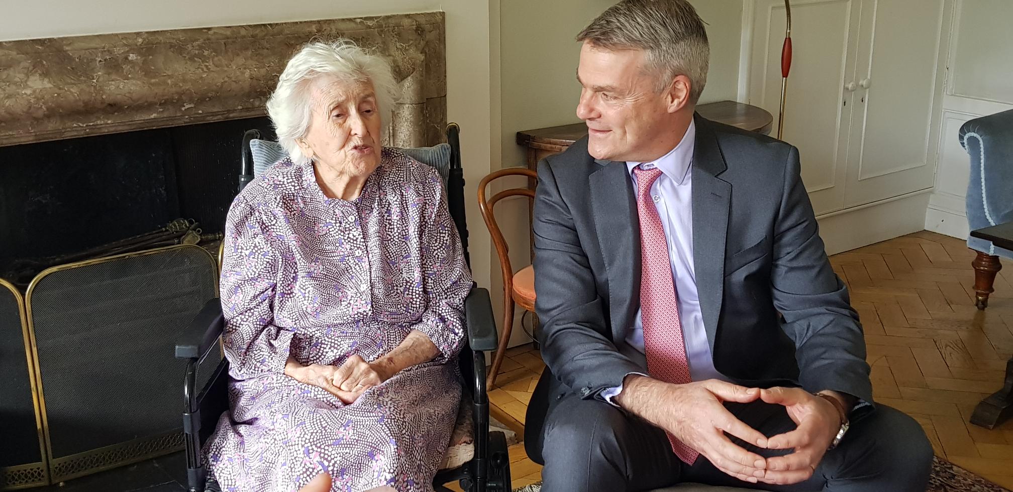 Elizabeth Kaser, who has short white hair and sits in a wheelchair, sits next to Professor Stephen Blyth, who wears a grey suit