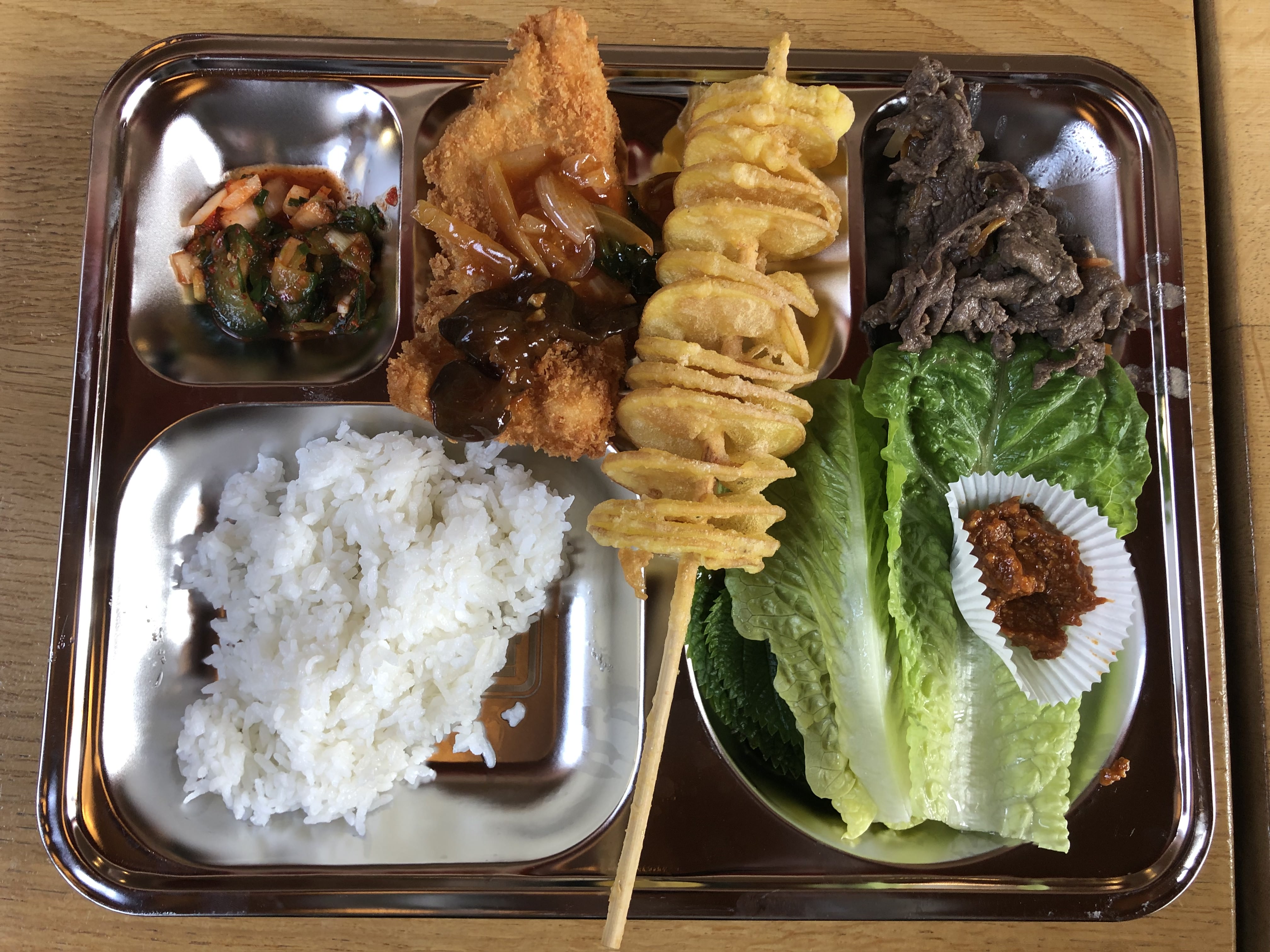 A shiny metal food tray with a selection of korean food, including rice, meat and a skewer with fried potatoes