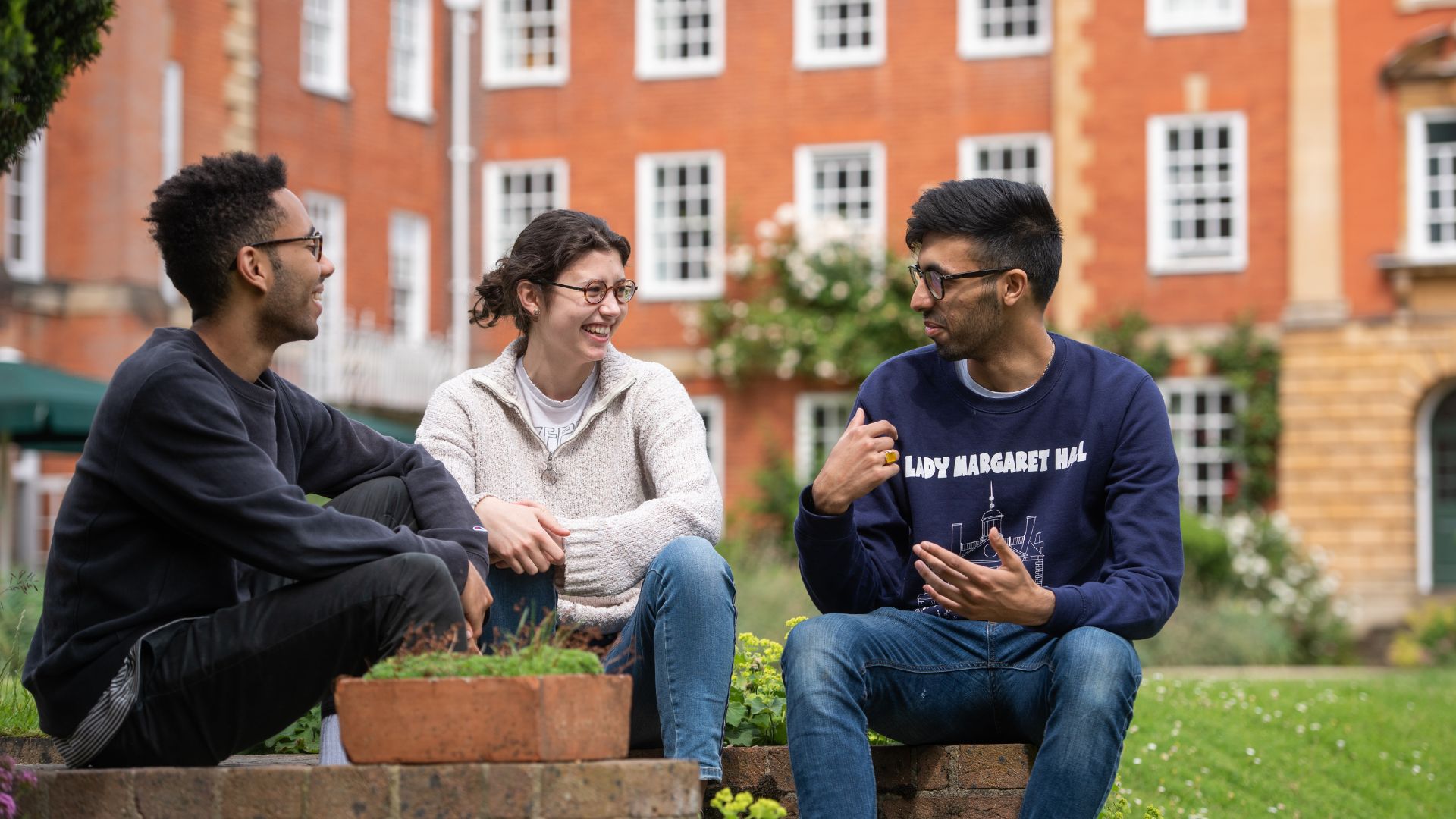 Three students, two men of Asian descent and a white woman, sit outside a red brick building talking and laughing