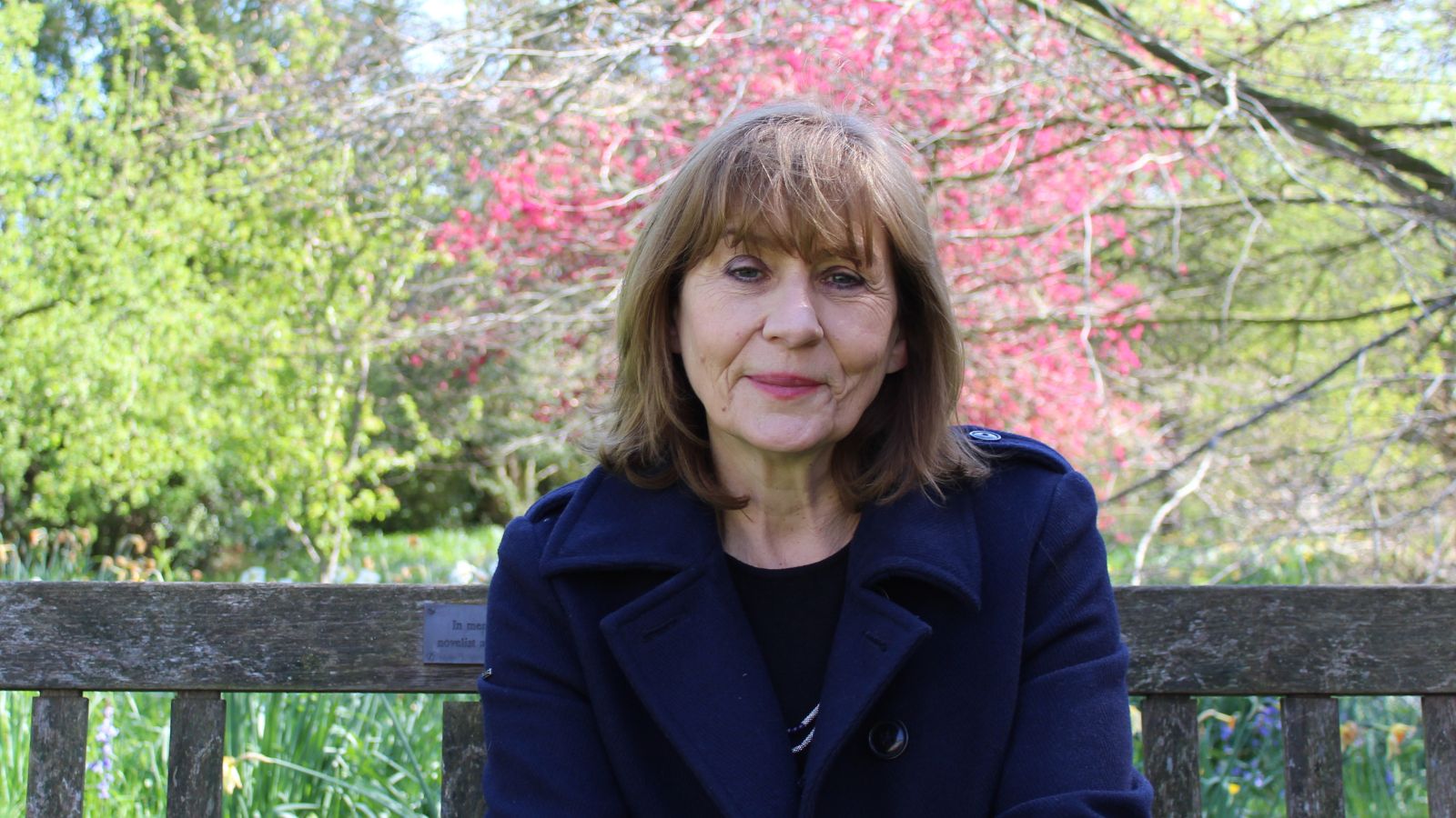 A woman with shoulder length dark brown hair wearing a dark blue coat sitting underneath a tree with pink blossom