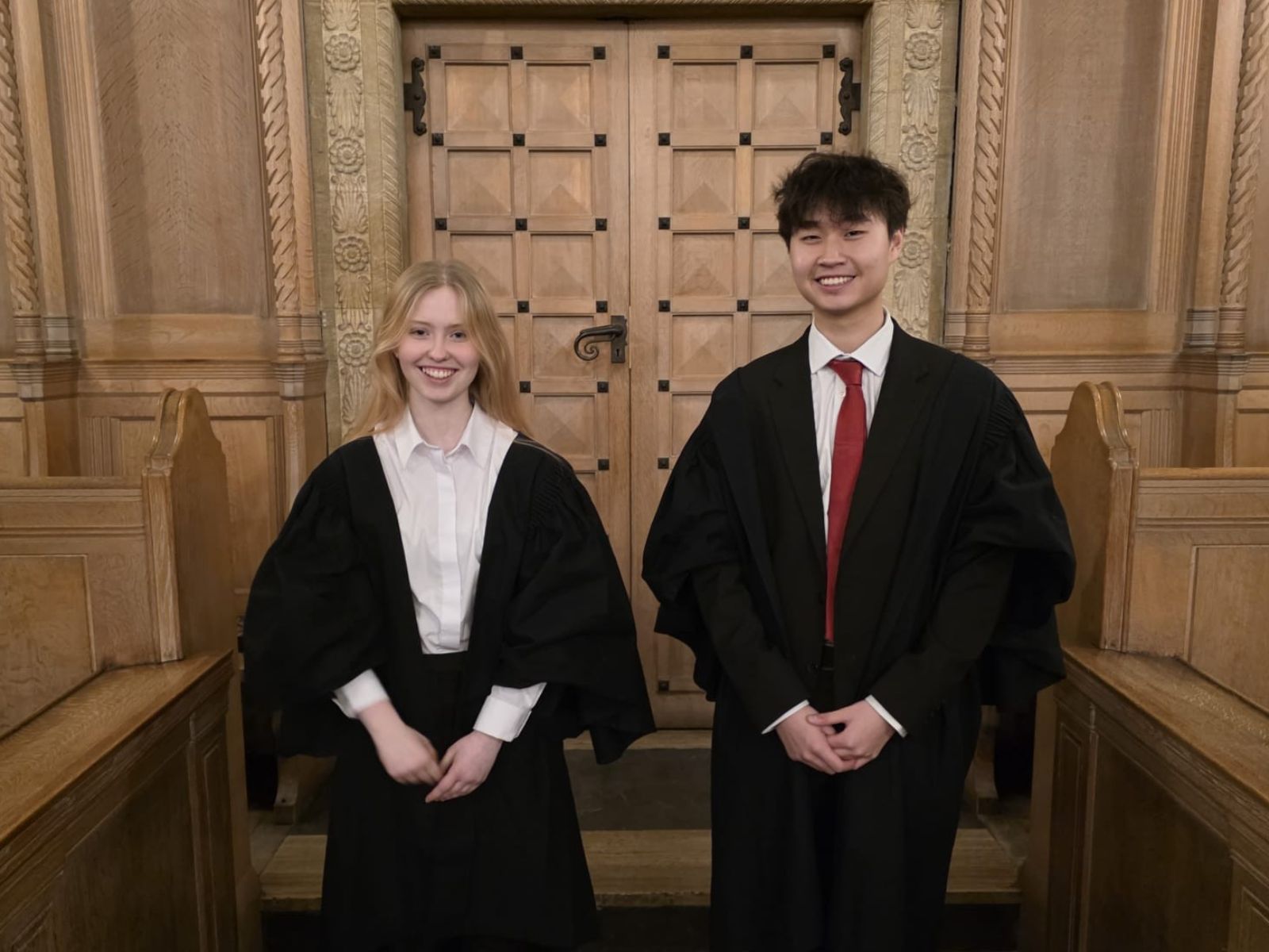 Two students in black academic robes stand in front of wooden doors in a Chapel