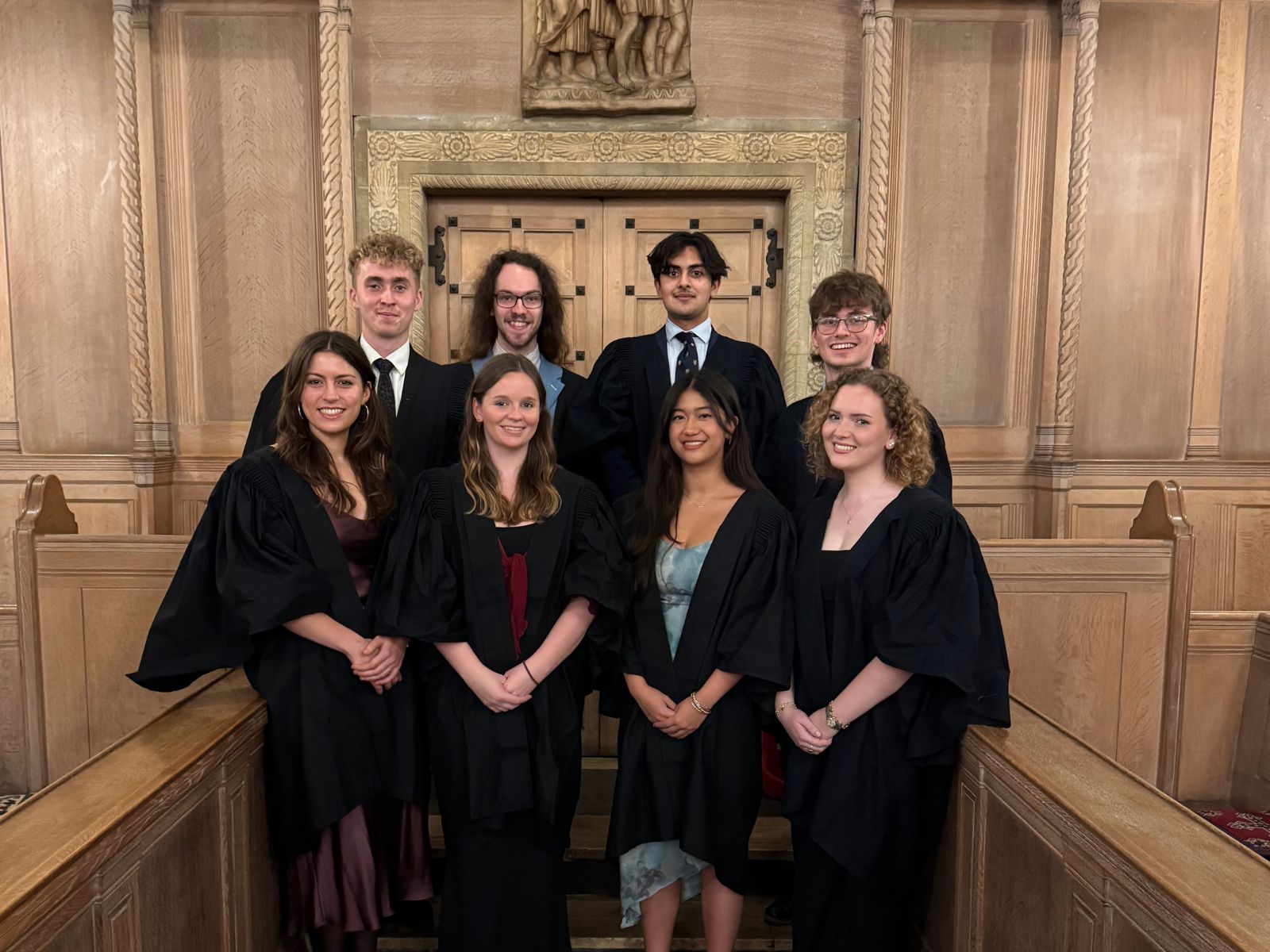 A group of students in black academic robes standing in front of ornately carved wooden doors