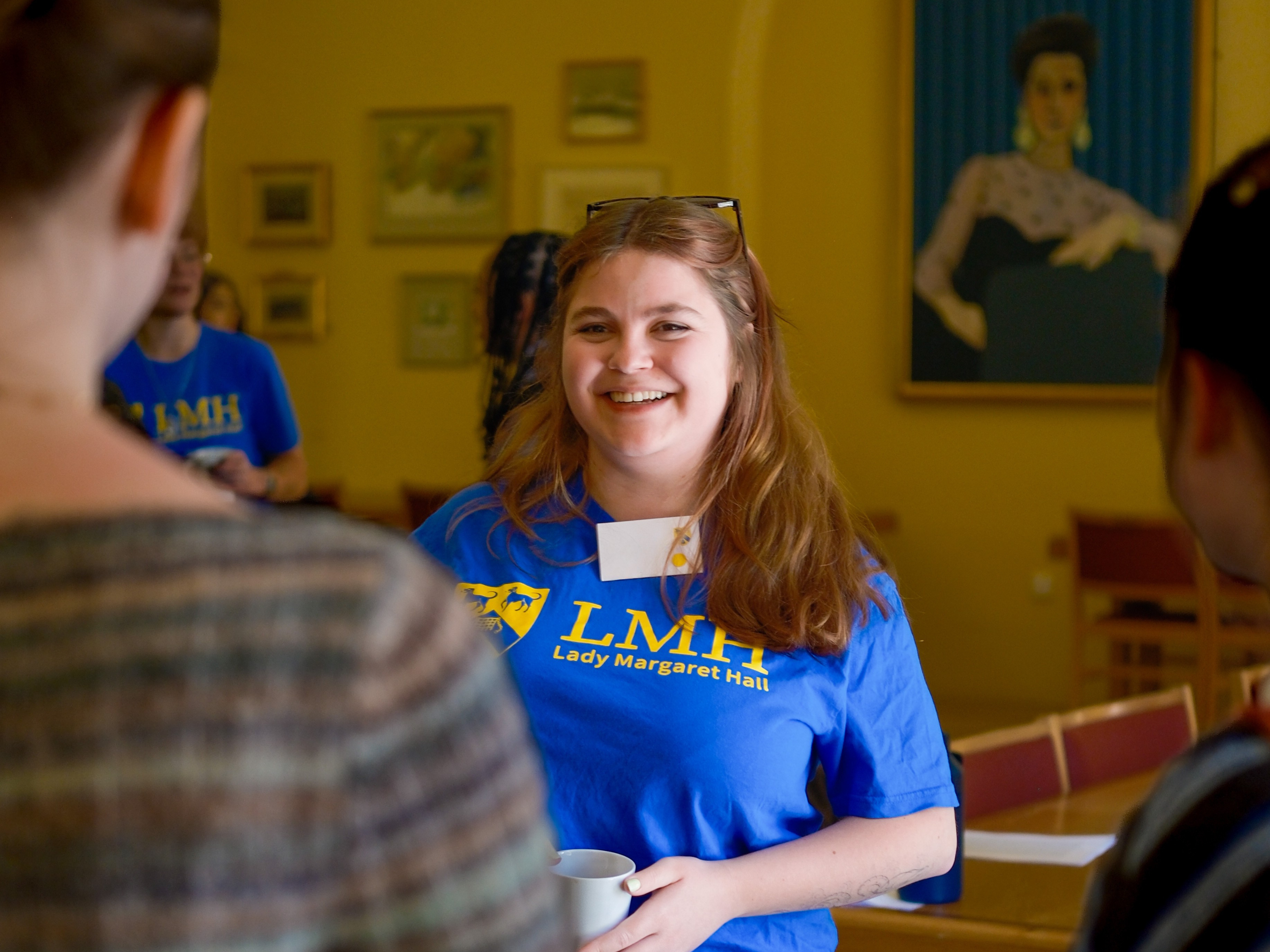A student ambassador wearing an LMH t-shirt smiles while chatting with prospective students at an outreach event. Other participants stand nearby in a warmly lit room with paintings on the walls, creating a welcoming and informal atmosphere.
