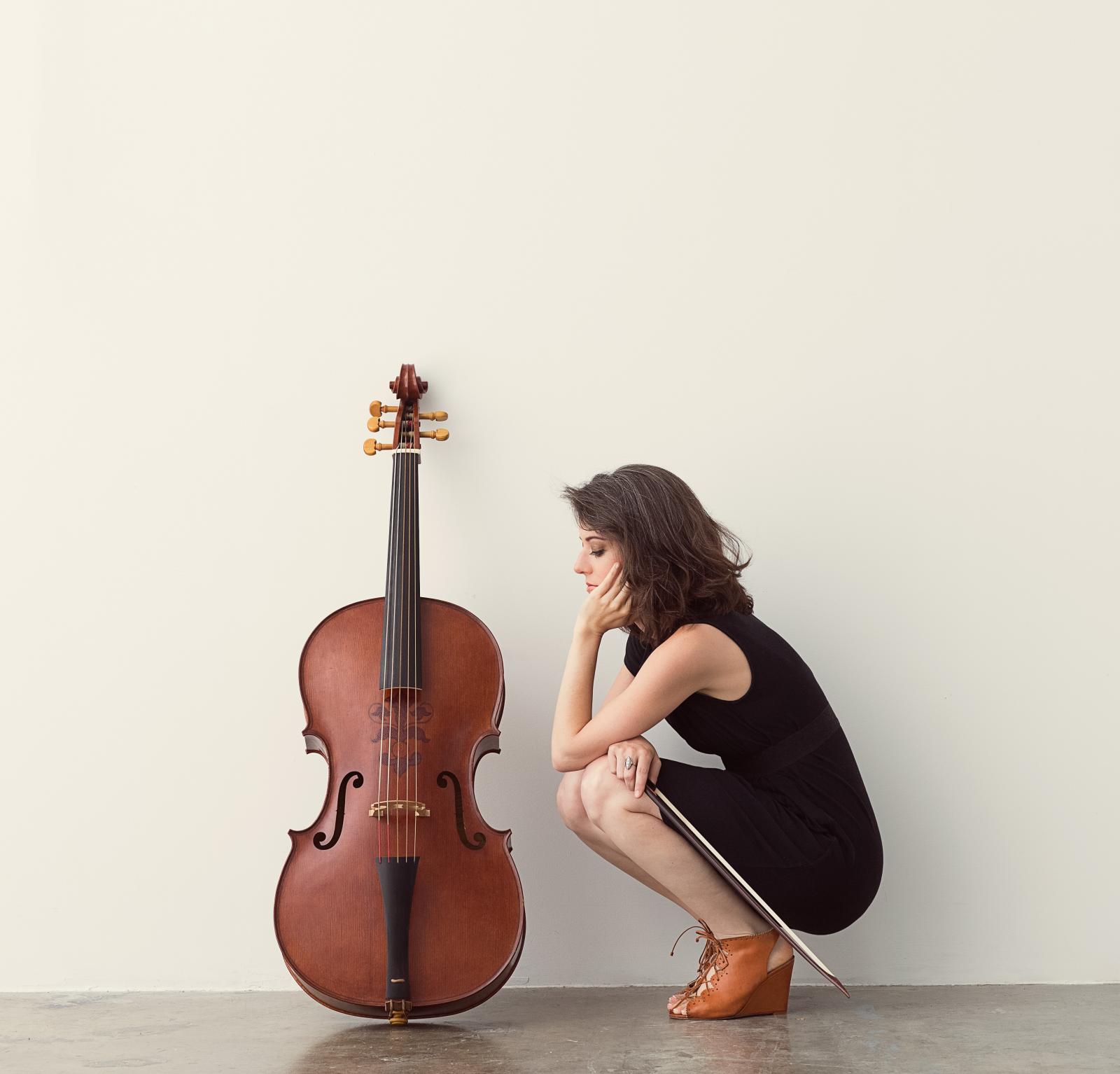 Elinor Frey with short brown hair in a black dress and brown shoes crouching by cello