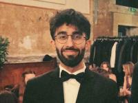 Photo of a young man with dark hair, a beard and glasses in a black suit