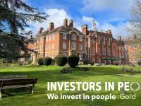 A red brick building set in gardens featuring green grass and large trees, with a blue sky