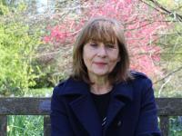 A woman with shoulder length dark brown hair wearing a dark blue coat sitting underneath a tree with pink blossom