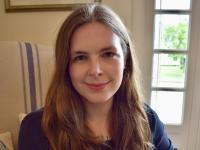Dr Grace Mallon, a woman with long brown hair, sits in a light-filled room