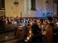 A candlelit carol service in the Lady Margaret Hall chapel. The choir is gathered in the front pews, holding lit candles and singing, while a speaker stands at the lectern. Congregants fill the pews, also holding candles. A lit Christmas tree glows to the right of the altar. The atmosphere is warm and reflective, with stained glass window reflections cast on the wall.