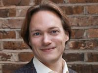 Tom Littler, who has mid-length red/brown hair and is wearing a grey suit with pale shirt