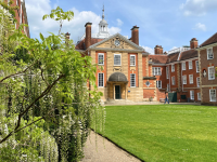 In the foreground, white wisteria is growing up the wall of the building. Further away is an imposing red brick building set in a quad