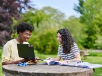 Two students sitting at a wooden table in the LMH gardens with books and laptops in front of them