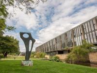 An abstract sculpture stands outside a 1960s modernist building with many windows, surrounded by green grass and trees