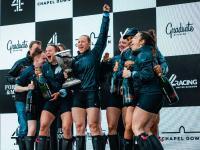 A group of 8 women and two men stand on a podium celebrating and holding a silver trophy after winning the Boat Race
