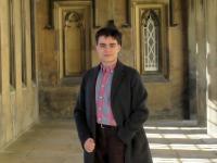 A young man in a long black coat standing in some stone cloisters