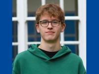 Jude Wood, who has short mid-brown hair and glasses. Jude is wearing a green hoodie and standing in front of a window