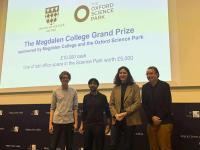 Four students stand on a stage in front of a screen saying: The Magdalen College Grand Prize