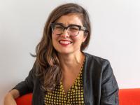 Professor Ana Domingos seated and smiling, wearing black-rimmed glasses, a black jacket, and a yellow and black polka-dot blouse, with a plain white wall in the background.