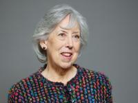 Baroness Manningham-Buller, an older woman with grey hair, wearing a colourful textured jacket, smiling against a plain grey background