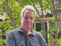 Professor Jochen Koenigsmann sitting outdoors in front of a stone building and leafy green plants, wearing a dark grey collared shirt