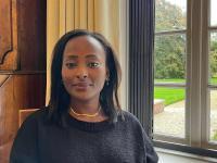 Dr. Makena Micheni sitting indoors by a large window with a garden view, wearing a black sweater and gold necklace, smiling softly at the camera