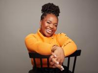 Dr Vee Kativhu sits on a backwards chair smiling expressively at the camera