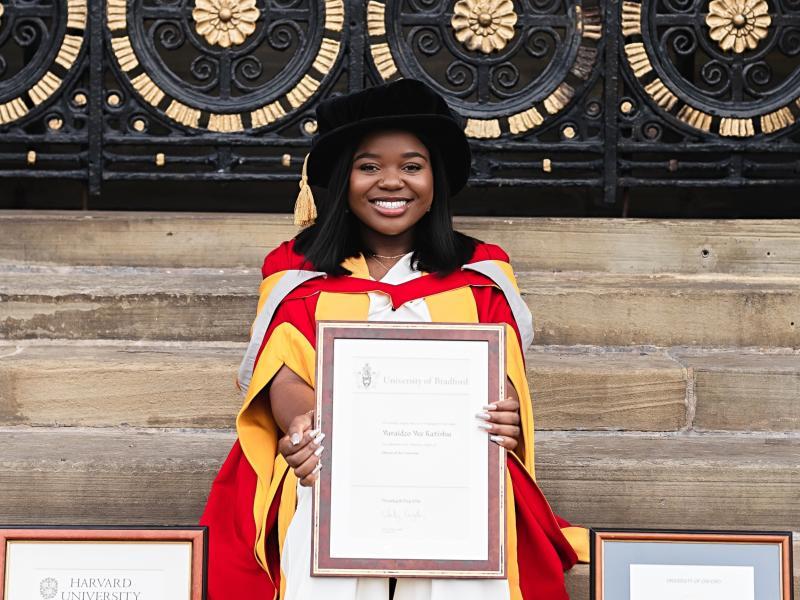 Vee Kativhu seated on some stone steps holding an honorary doctorate from the University of Bradford