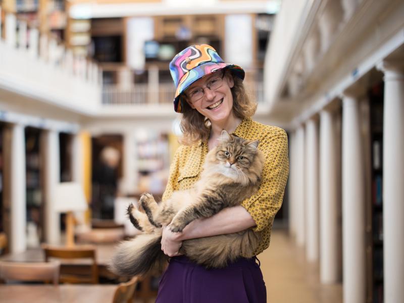 College Librarian Jamie Fishwick-Ford and their cat Isambard standing in the LMH Library