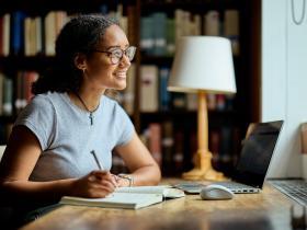 A young woman with tied-back curly brown hair sits at a desk in a library with her laptop