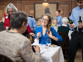 A lady in a blue top smiles while chatting with guests at a formal LMH event in a wood-panelled dining hall. Around her, alumni, supporters and students are seated at tables in conversation, while student servers carry bottles of wine through the room. The atmosphere is warm, lively and sociable.