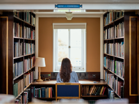 A student sits at a desk in a quiet library alcove, facing a window framed by bookshelves filled with books. Natural light streams in, creating a calm and focused study environment.