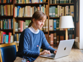 A student sits at a wooden table in a library, working on a laptop. Behind them are tall bookshelves filled with books, creating a warm academic setting. She is wearing a blue jumper over a light-coloured collared top and appear focused on the screen. A table lamp stands nearby, and daylight enters from a window on the right. The scene conveys quiet study or research in a library environment.