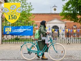 A woman pushes her bicycle past the gates of Lady Margaret Hall