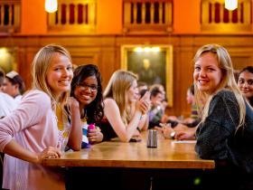 A group fo female students smile to the camera while sitting around a dining hall table.