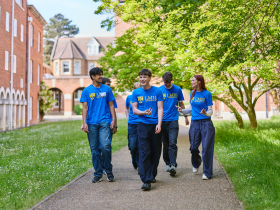 A group of student ambassadors wearing LMH t-shirts walk together along a tree-lined path on campus. Surrounded by red-brick college buildings and green lawns, they chat and laugh as they make their way through the College grounds.