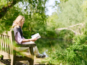 A student sits on a wooden bench beside a river, reading a book in the sunshine. Surrounded by lush green trees and foliage, she enjoys a peaceful moment outdoors in a tranquil garden setting.