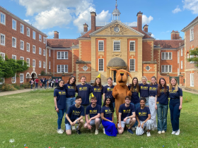 A group of smiling students wearing matching navy blue LMH (Lady Margaret Hall) t-shirts pose in front of a grand brick college building with a domed entrance. A person in a large brown dog mascot costume stands at the center. The group is gathered on a green lawn under a sunny sky with fluffy clouds.