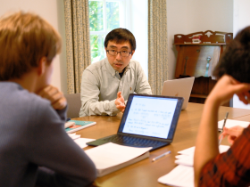 An LMH Fellow leads a small-group tutorial discussion around a table, speaking to students seated with notebooks and laptops in a bright study room. The students listen attentively.
