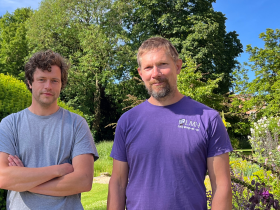 Two men in casual t-shirts standing in some gardens with blue sky and trees in the background