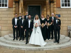 A wedding party stands on the steps to Talbot Hall, with the bride wearing a long white dress and everyone else in darker formal outfits