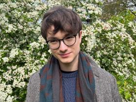 Leo Gitin, who has short dark brown hair and glasses, standing in front of a bush with lots of small white flowers
