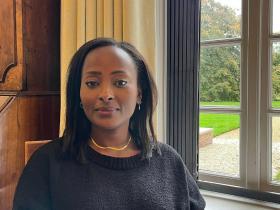 Dr. Makena Micheni sitting indoors by a large window with a garden view, wearing a black sweater and gold necklace, smiling softly at the camera