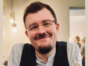 Florian Lutzelberger, who has short dark hair and a dark beard and moustache and clear-framed glasses is smiling at the camera