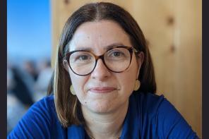 Maria Coelho, who has short dark brown hair and glasses and is wearing a blue top, smiles at the camera