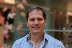 Oliver Pambos, who is wearing a pale blue shirt and standing in an atrium