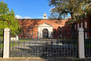 A beautiful image of the front of Lady Margaret Hall, with a set of wrought iron gates in front of an imposing red brick building set against a bright blue sky