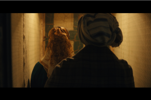A still from Onyeka Igwe's film 'A Radical Duet', showing two women wearing headscarves facing away from the camera and walking down a dimly lit corridor