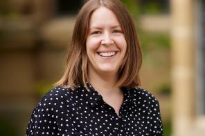 A smiling woman with long brown hair wearing a black and white polka dot dress standing in front of a stone building